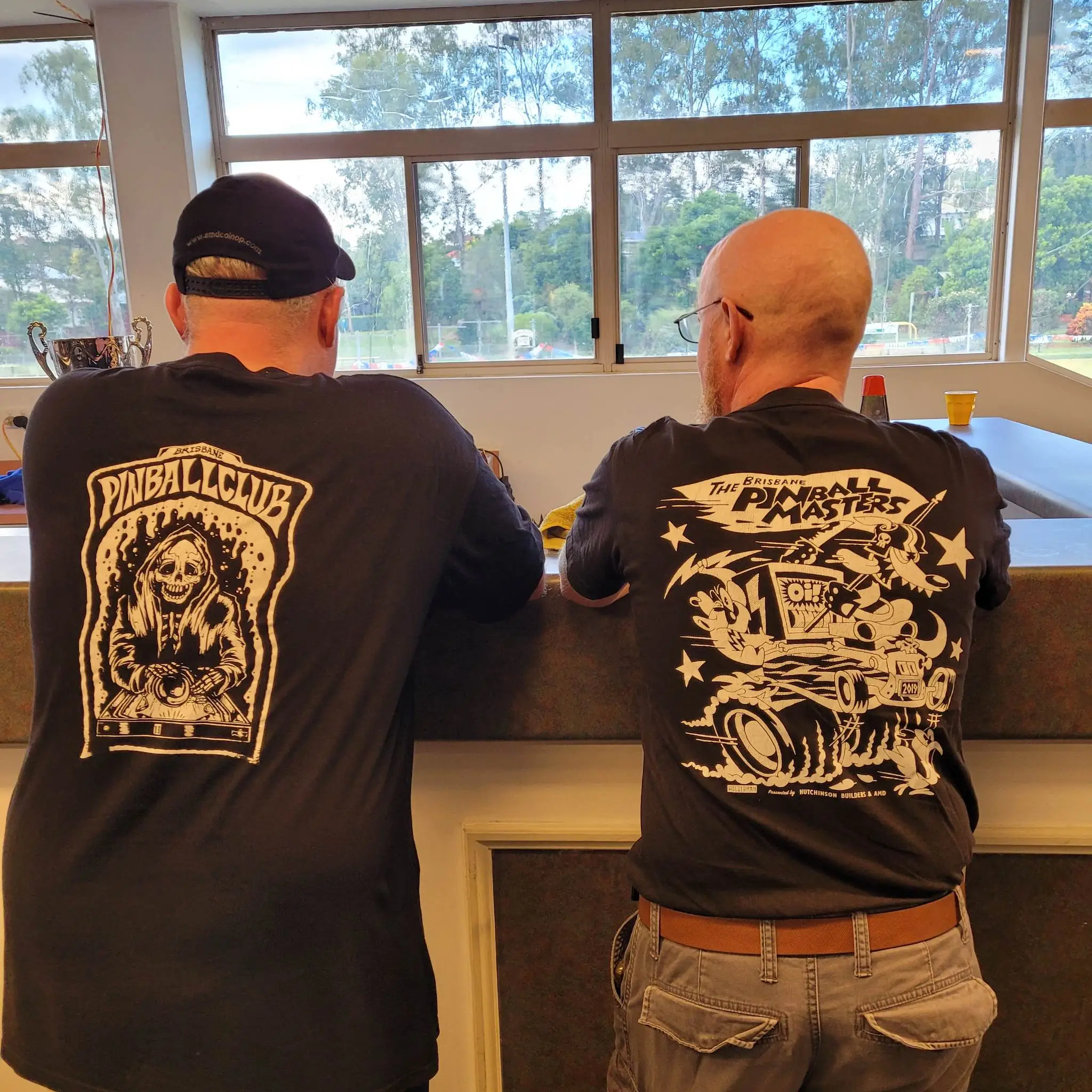 Two pinball enthusiasts in matching shirts look out window at trees